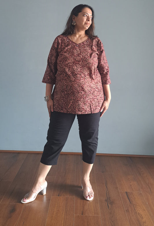 Woman wearing a kalamkari top and black pants standing on a wooden floor with a gray wall background